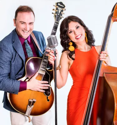 Woman in orange dress holding a cello and a man in a blue dress jacket holding a guitar.