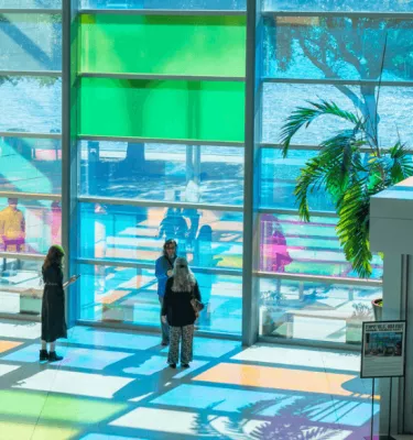 People standing around a room filled with glass windows that are letting lights of green, blue and pink in.