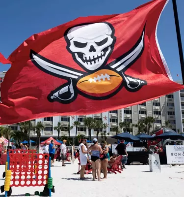 A group of people standing on a beach playing games and mingling with the Tampa Bay Bucs red flag blowing in the wind.