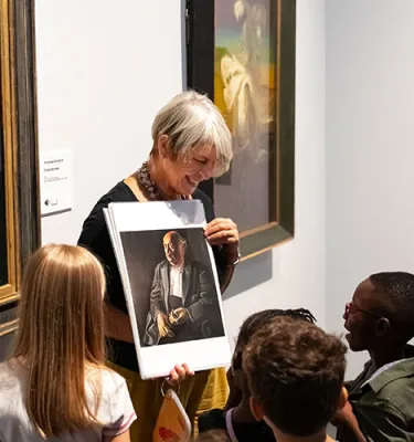 Older woman with short hair showing a group of young kids of painting photo inside a museum.