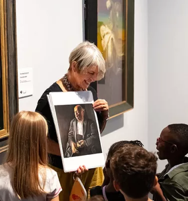 Older woman with short hair showing a group of young kids of painting photo inside a museum.