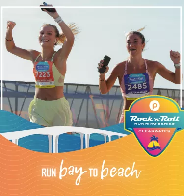 Two women runners in a race outside during the day with their arms up and smiling.