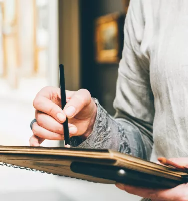 Photo of a hand holding a black pen sketching on a brown notebook.
