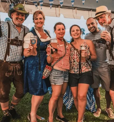 Six adults dressed in Octoberfest outfits holding beers and smiling at the camera.