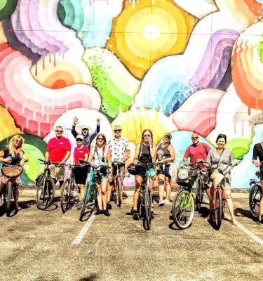 A group of adults on bikes in front of a colorful wall mural and smiling at the camera.