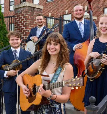 Group shot of young adults holding string instruments on outside steps and smiling at the camera.