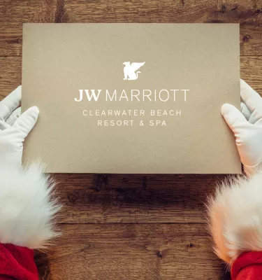 Hands of Santa Claus holding a light brown box that says "JW Marriott Clearwater Beach Resort & Spa."