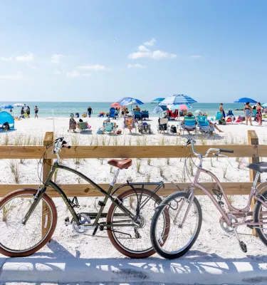 two bikes lean up against a wooden fence near a white-sand beach with umbrellas and people in chairs