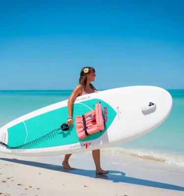Woman walks along the ocean holding a stand-up paddleboard under her arm.
