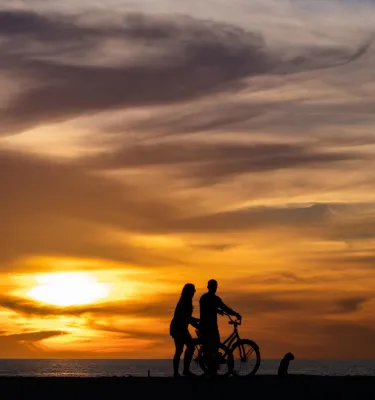 a dramatic orange sunset backlights two people with a bike on the beach at Treasure Island