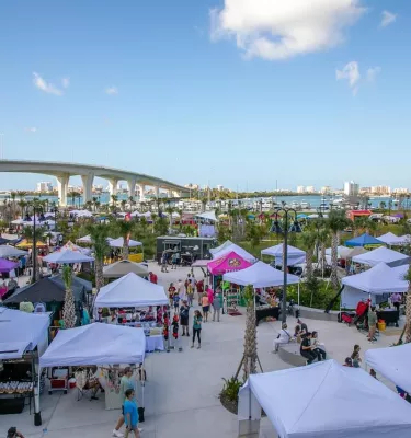 Aerial view of an outdoor market on the water featuring multiple vendor tents and crowds of people.