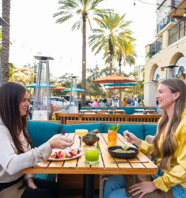 Two women diners enjoy food and drinks at an outdoor restaurant with palm trees in the background
