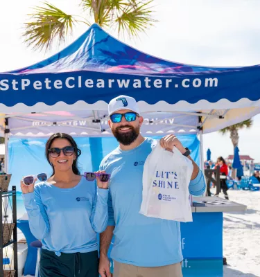 two people in St. Pete/Clearwater blue shirts hold up sunglasses and a swag bag at an event on the beach