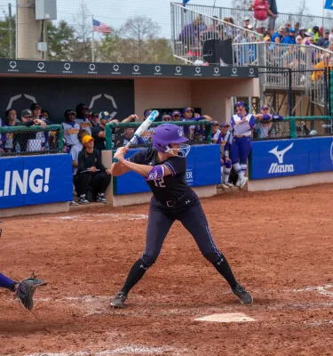 a young woman at bat with a catcher and umpire behind her at a softball tournament with crowds in the stands