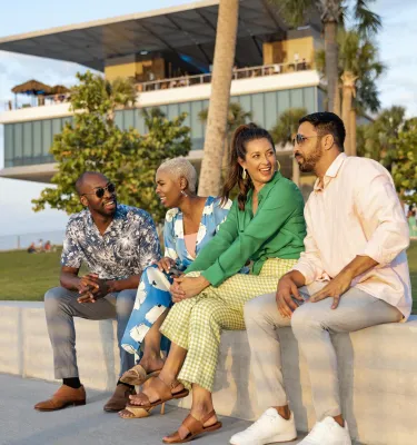 four people sit on a short wall talking with the St. Pete Pier in the background