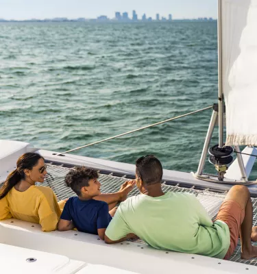 a family on the netting of a sailing catamaran on the water with buildings in the distance