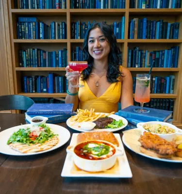 A woman in a yellow dress holds up a cocktail in front of plates of food with a bookshelf behind her