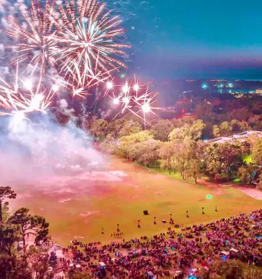 Independence Day fireworks in a park with a crowd of people watching during the evening.