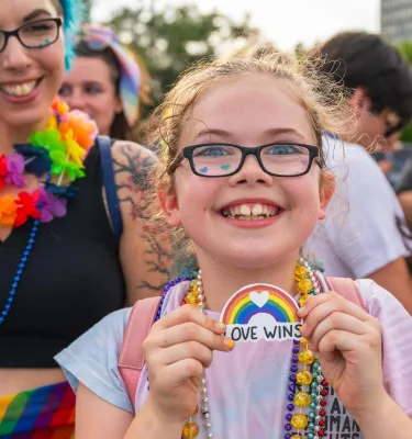 a smiling girl wearing glasses and rainbow beads holds a "love wins" rainbow sign while standing next to her mom