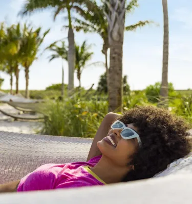 Woman with sunglasses laying in a hammock with the beach and palm trees in the background