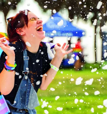 a person smiles joyfully while wearing a rainbow cape at an outdoor pride event