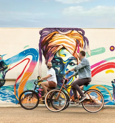 two men riding bikes in front of a colorful mural of an african woman wearing a turban