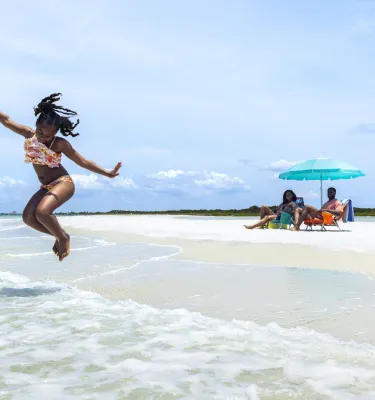 a little girl in a pink bathing suit jumping at the water's edge at the beach while her parents relax under an umbrella