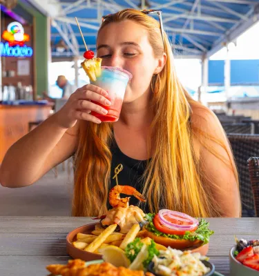 a woman sipping a red white and blue frosty cocktail at an outdoor restaurant with a sandwich and fries in front of her