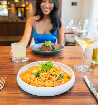 a smiling woman in a bright blue dress sits across from a bowl of pasta at a restaurant