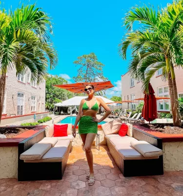 Una mujer con un traje de baño verde brillante posando frente a bancos y una piscina cerca de palmeras en el Hotel Fenway