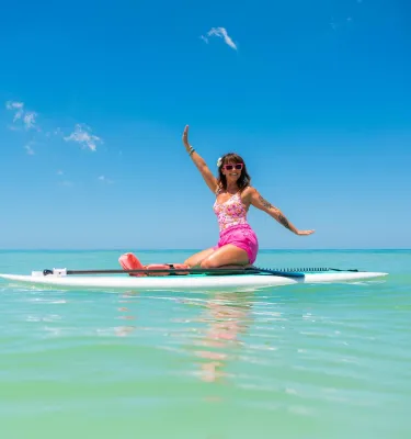 a woman dressed in pink clothing and sunglasses smiles as she sits on top of a paddleboard at the beach