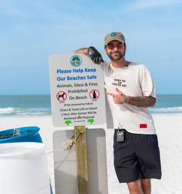 a young man standing next to a trash can and a sign at the beach encouraging people to pick up trash