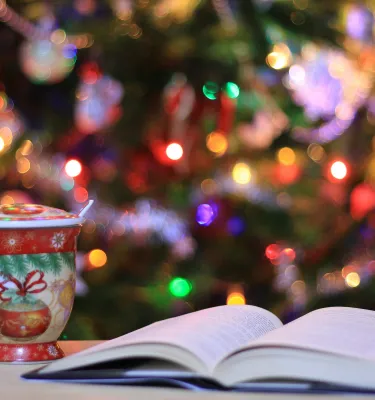 An open book on a counter with a tea mug and a Christmas tree lit up in the background.