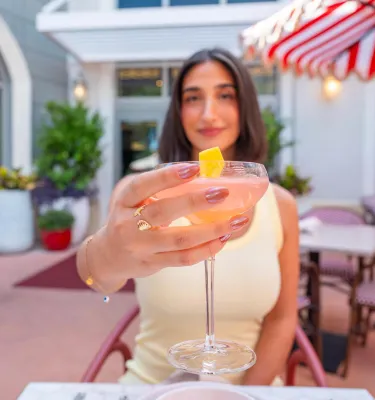 a woman holding out a cocktail toward the camera; she is sitting an an outdoor table near a striped umbrella at Juno & The Peacock