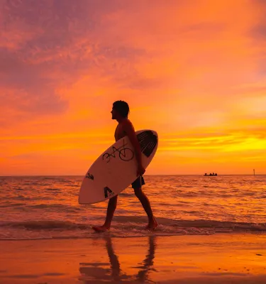 a man with a surfboard walks along the water's edge in Treasure Island at sunset
