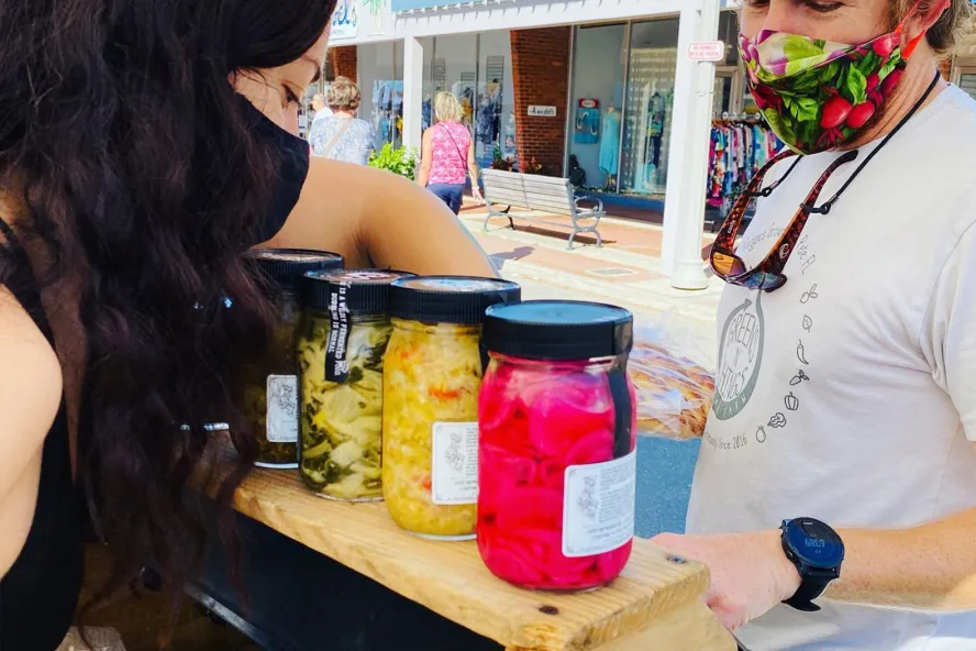 Uma mulher está vendendo grandes potes de legumes em conserva no Corey Avenue Sunday Market em St. Pete Beach.