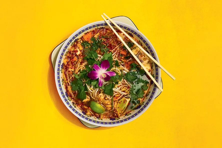 A Thai-soup plate with a purple flower on top of a yellow table. 