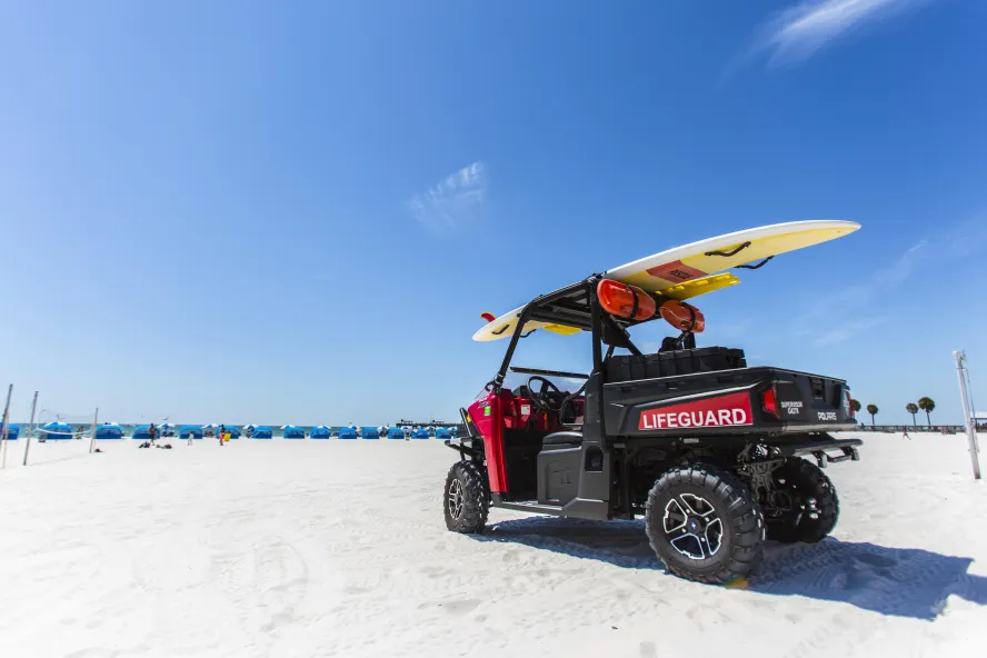 Am Strand parkt ein Rettungsschwimmer-Buggy mit einem Surfbrett darauf. In der Ferne sind Cabanas zu sehen.