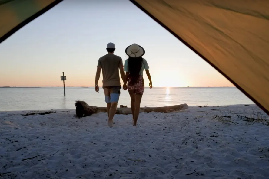 A couple walks away from a tent toward the water at Shell Key during sunset