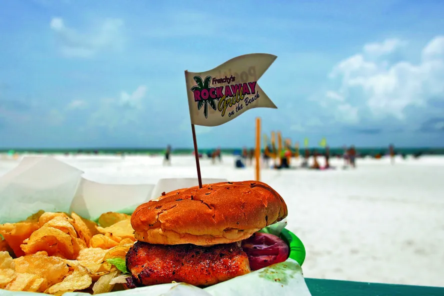 Ein Zackenbarsch-Sandwich mit Pommes auf einer Außenterrasse mit Blick auf den Strand. Strandbesucher sind schon von weitem zu erkennen.