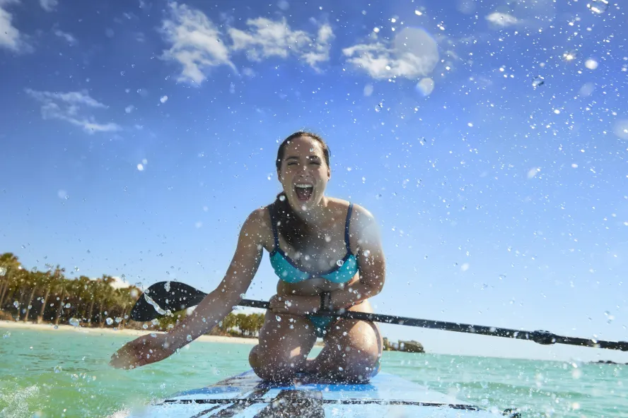 A girl splashing water at the camera while kneeling down on a stand up paddleboard.