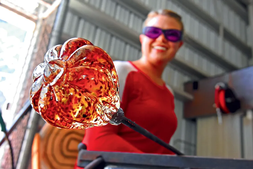 A female glass artist shows off a molten, orange glass art piece in progress at the Morean Arts Center's hot shop in St. Pete