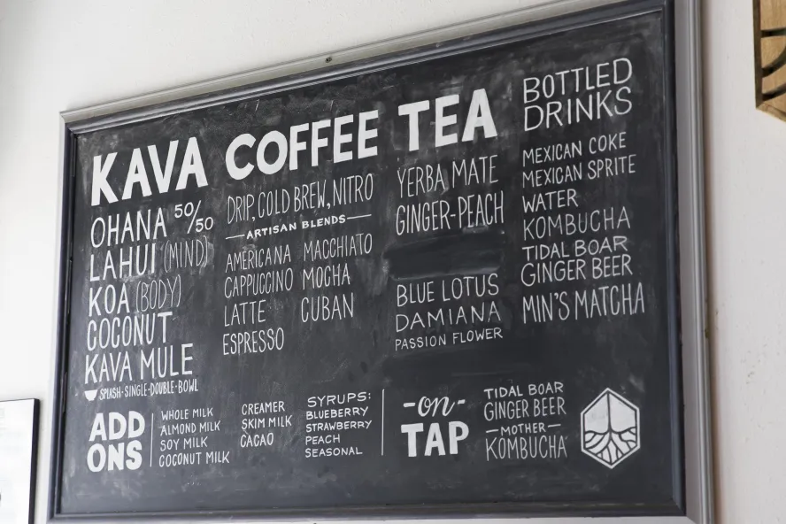 Imagen de una pizarra negra en una pared con KAVA COFFEE TEA en negrita y un menú que llena el resto de la pizarra contra una pared blanca.