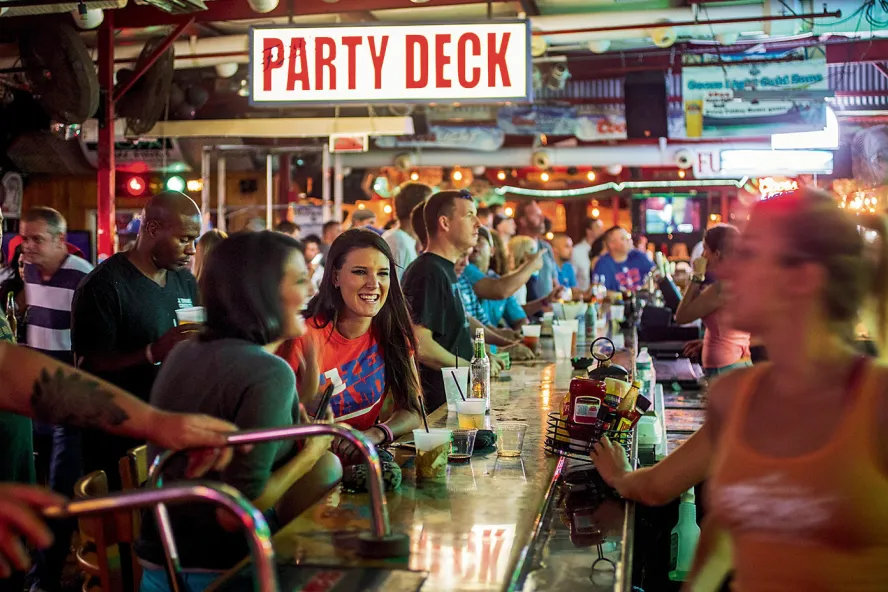Mucha gente sentada y de pie en un bar viendo deportes y bebiendo, un gran cartel en la parte superior de las imágenes que dice "Party Deck".