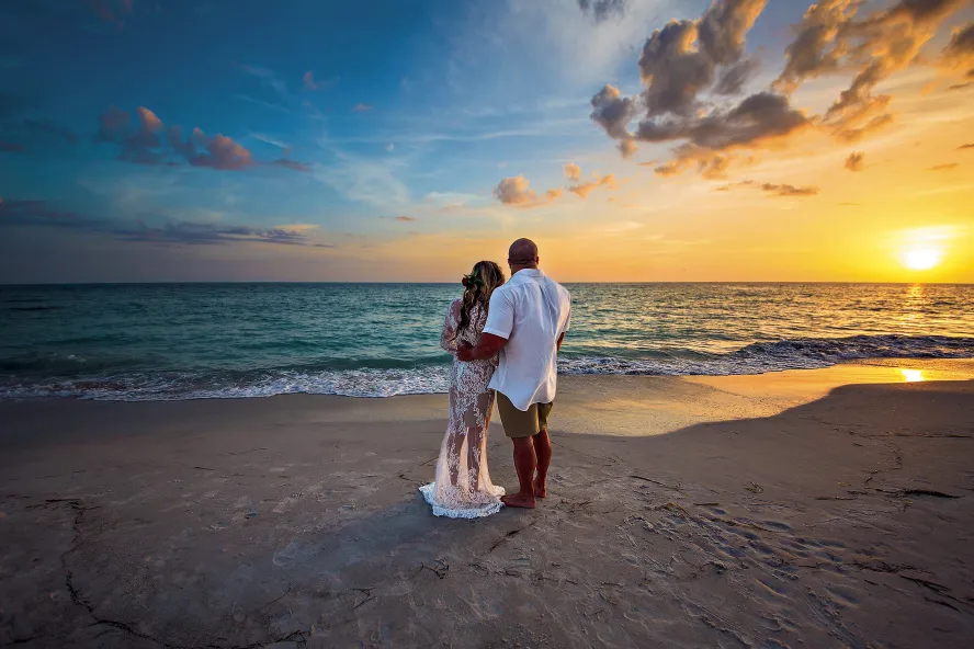 Bride and groom hugging each other and watching the sunset at the beach.