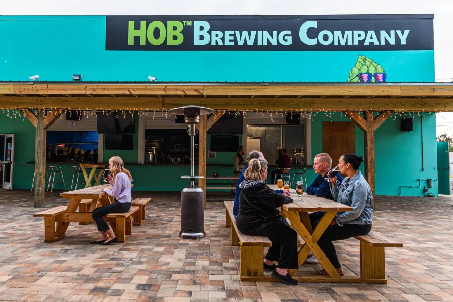 Several people enjoying beers outside of the HOB Brewing Co., seating on wooden benches.