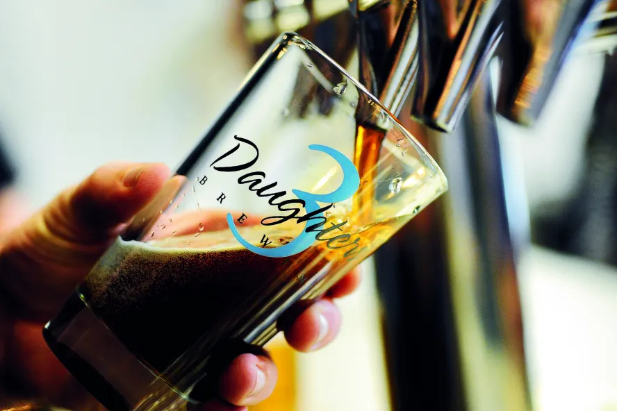 A person pouring dark beer in a 3 Daughters Brewery glass.