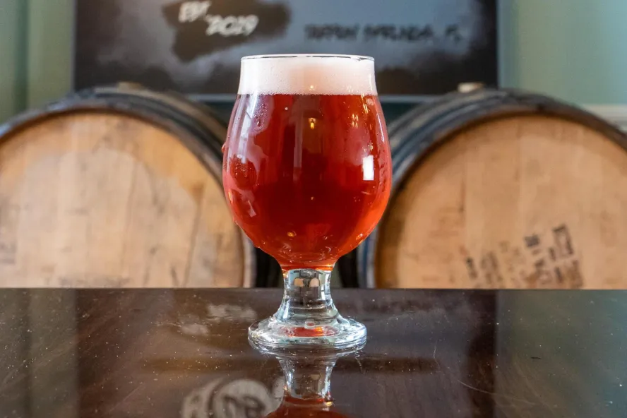 A round glass with red ale in front of wooden barrels.