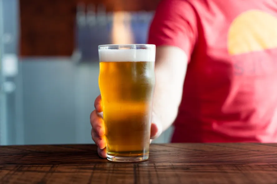 A person wearing a red shirt holds a glass of beer on top of a wooden table.