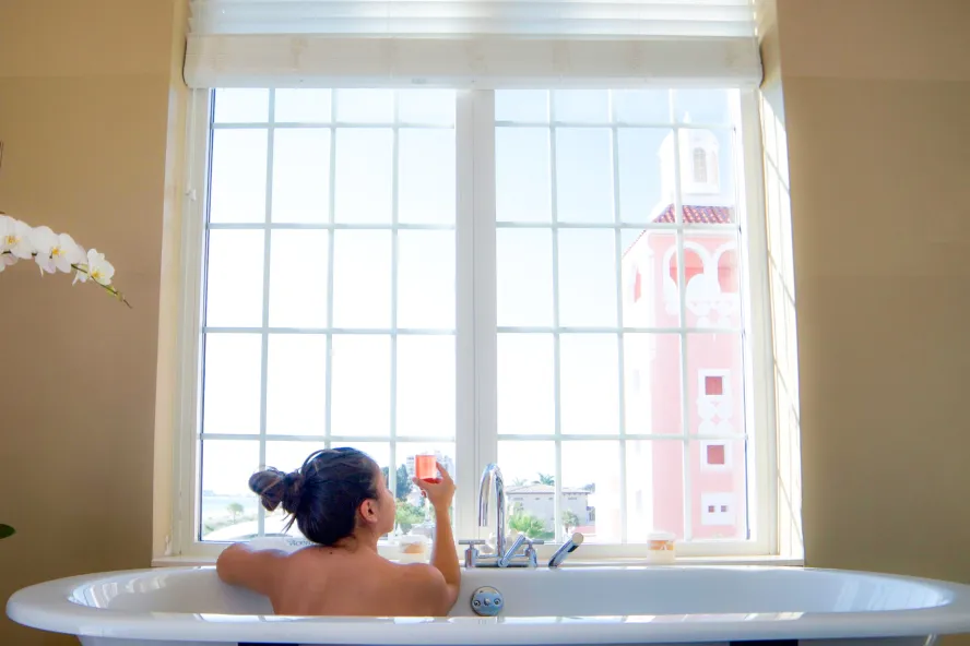 A woman in a bath tub holds a pink champagne glass while looking out the window at the Don Cesar at Spa Oceana